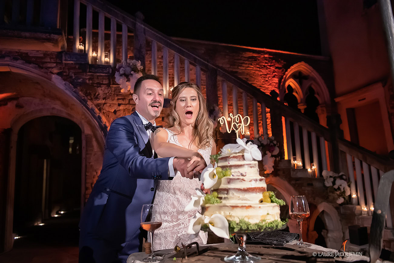 Joyful newlyweds cut their wedding cake in a romantic Venetian setting illuminated by warm, ambient lighting.