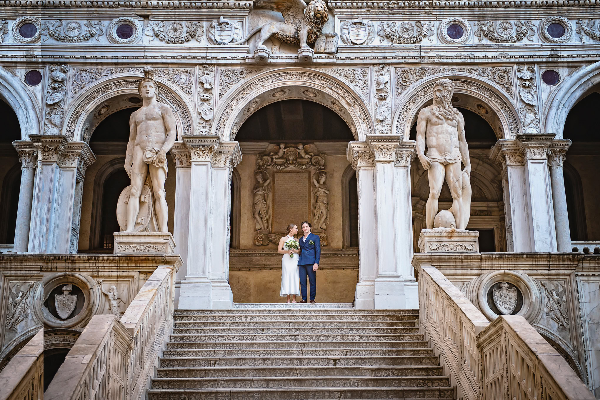 Elegant wedding couple standing on grand marble staircase beneath historic Venetian architecture and statues, bathed in.