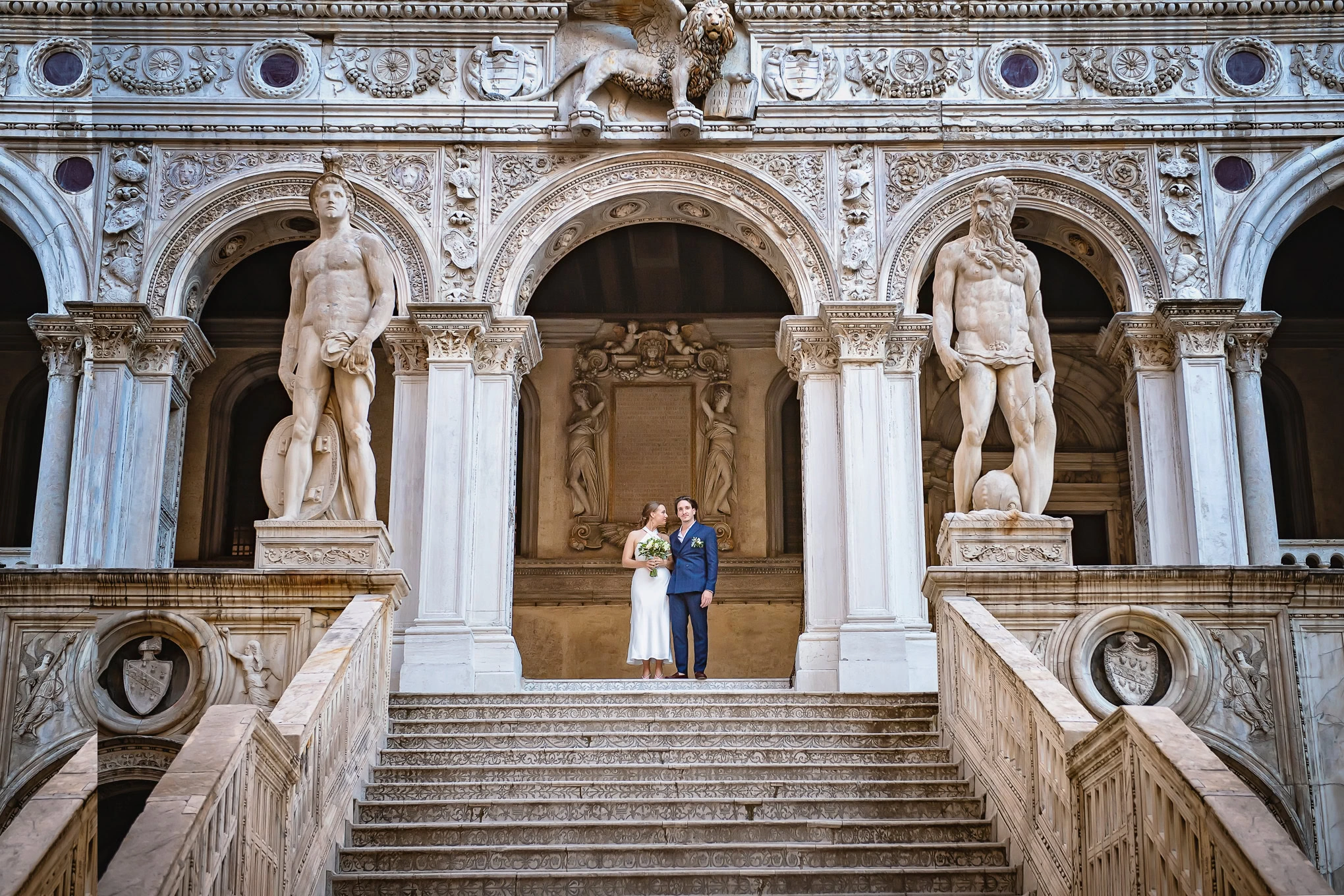 Elegant wedding couple standing on grand marble staircase beneath historic Venetian architecture and statues, bathed in.