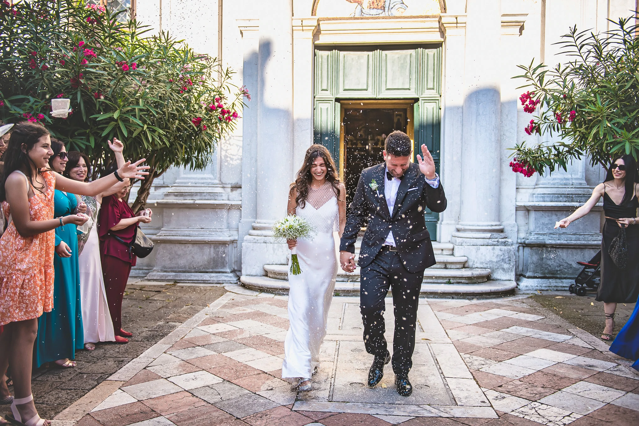 A couple in elegant wedding attire walking hand in hand out of a historic church in Venice, surrounded by joyful guests.