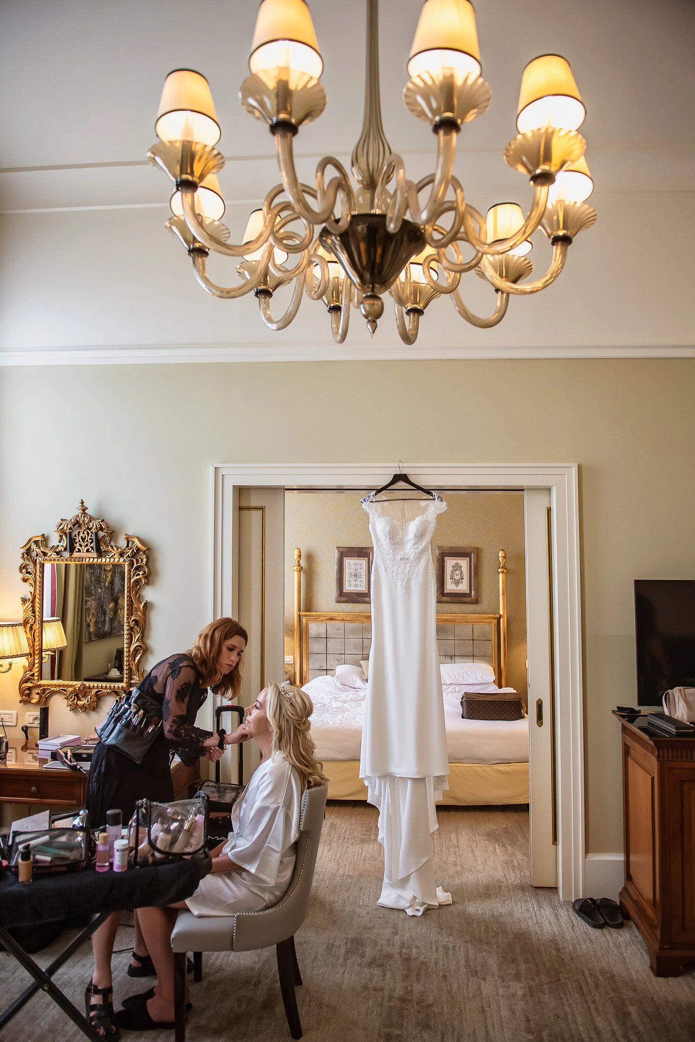 A bride getting ready in a luxurious Venetian hotel room, illuminated by warm chandelier light, with her wedding dress h.