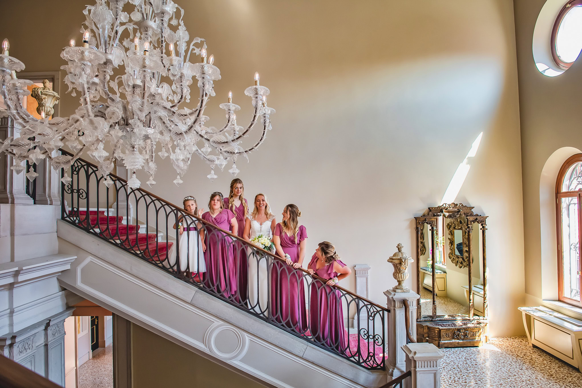 A woman and girls in pink dresses walking down a grand Venetian staircase with a chandelier overhead.