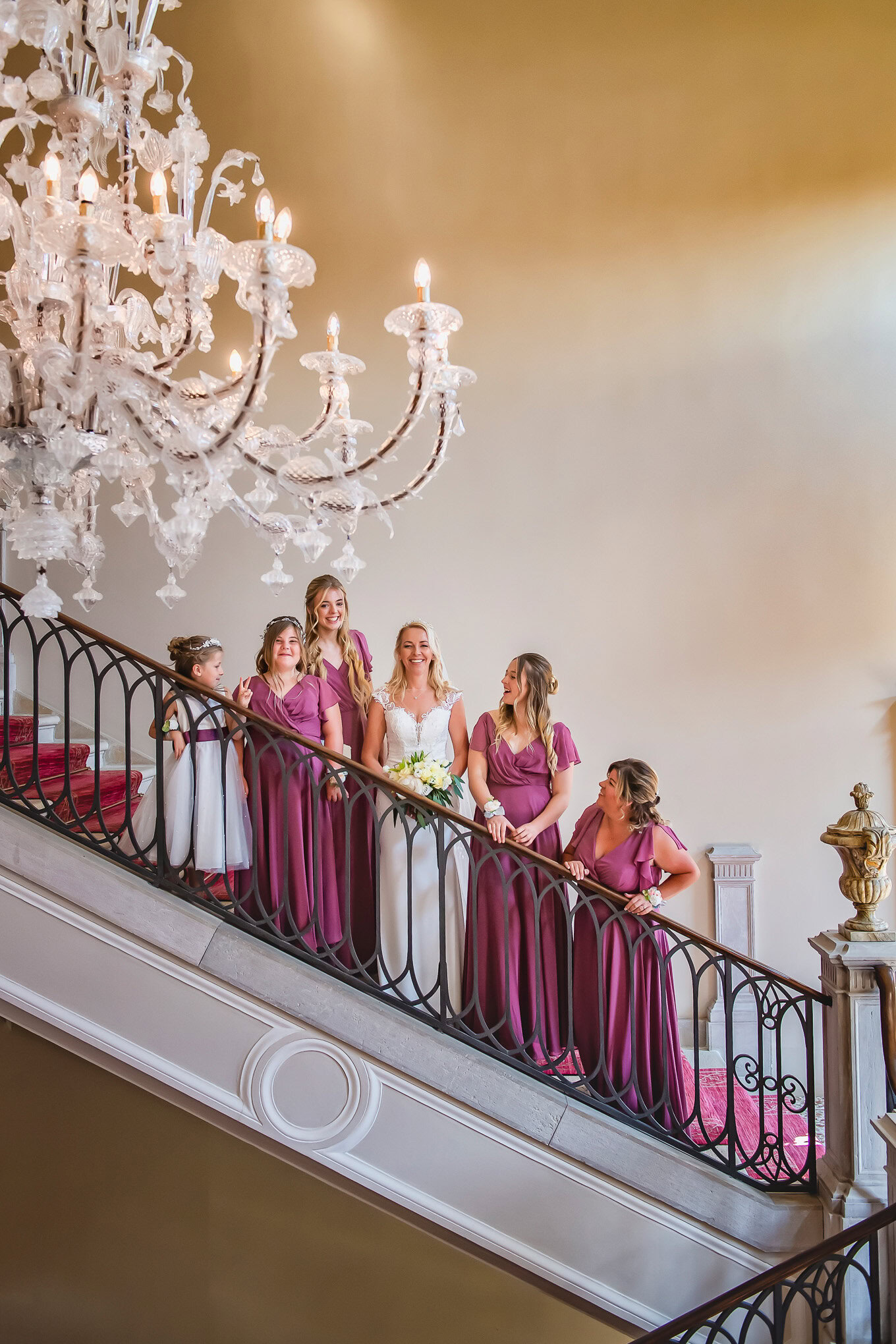 A bride and her bridesmaids in elegant purple dresses gather on a grand staircase illuminated by a sparkling crystal cha.