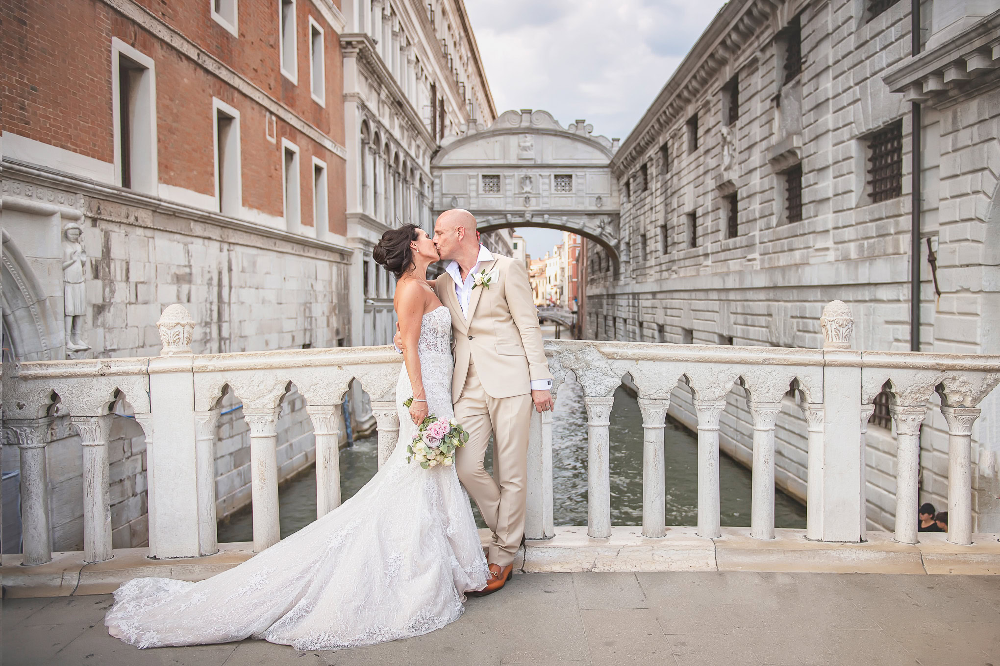 Romantic wedding moment in Venice with couple sharing a kiss on a historic bridge under soft daylight.