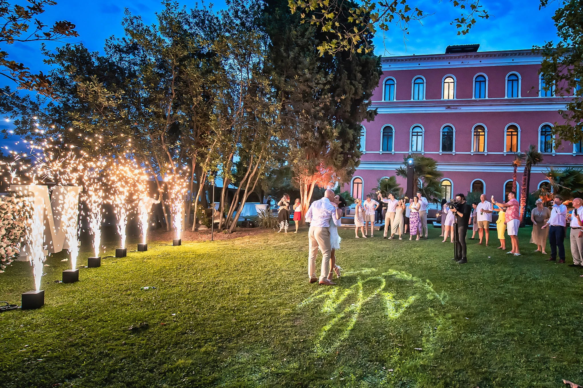 Elegant wedding celebration in Venice with fireworks, guests dancing on lush green lawn under twilight sky and historic.