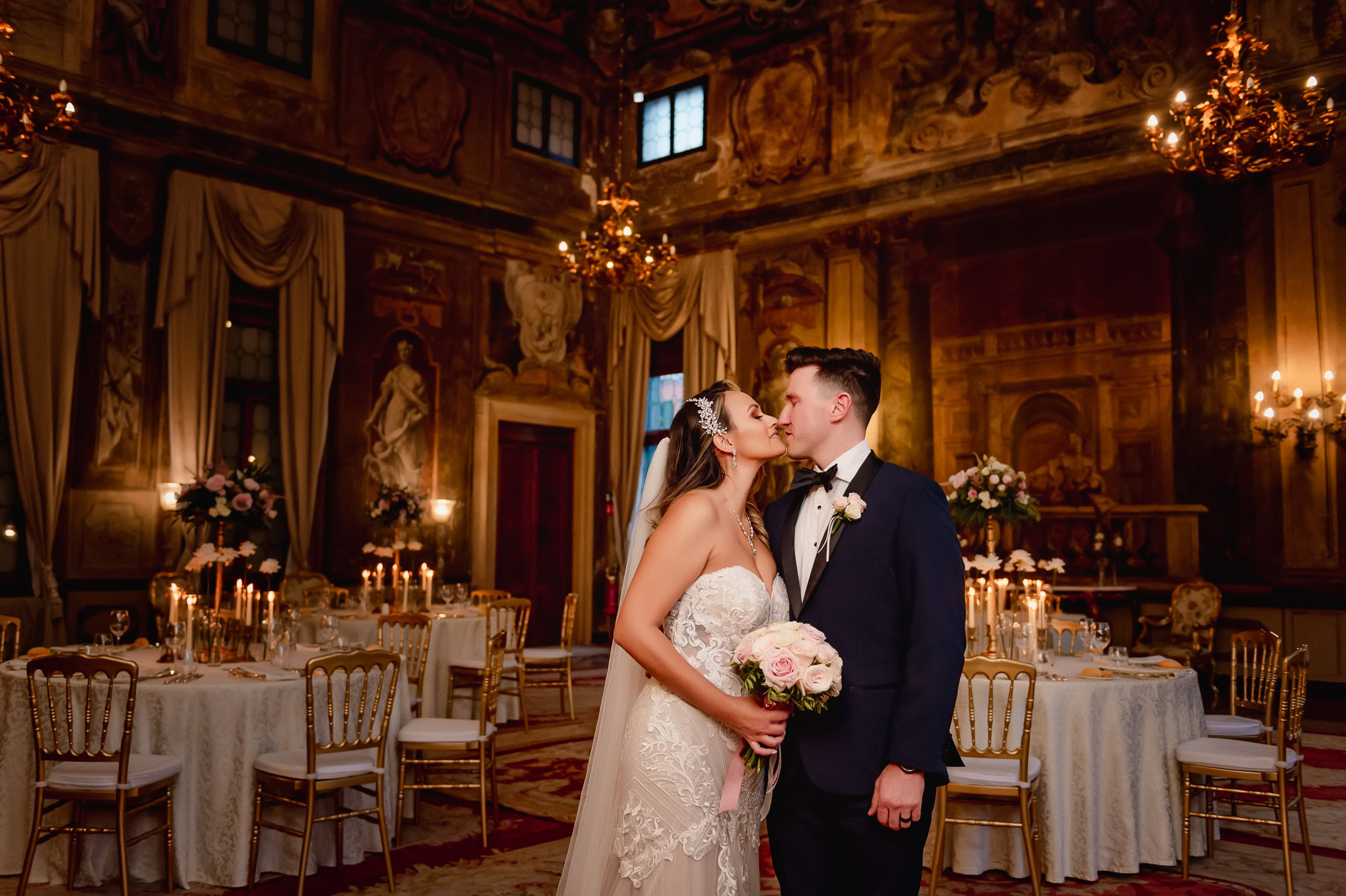 A beautiful bride and groom share a romantic kiss in an opulent Venetian ballroom illuminated by warm candlelight.