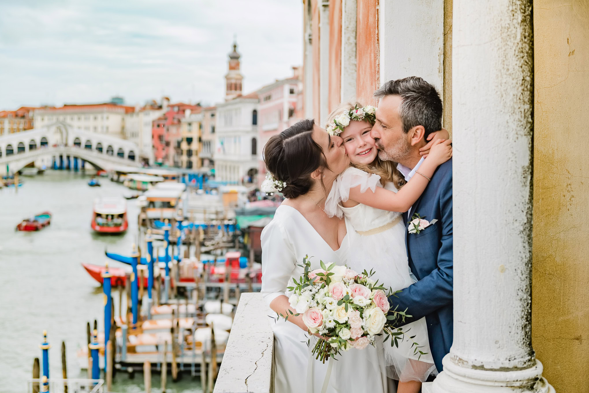 Romantic wedding moment in Venice with a couple sharing a kiss and a young girl, surrounded by colorful buildings and th.