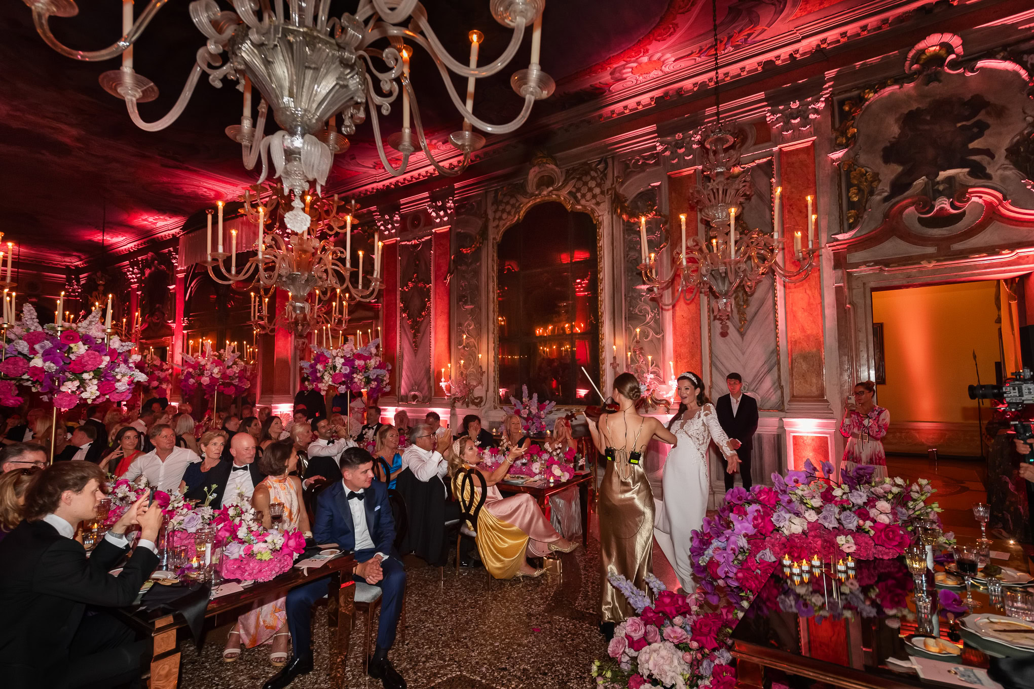A wedding couple dances gracefully in an opulent Venetian ballroom illuminated by warm pink and gold lighting, surrounde.