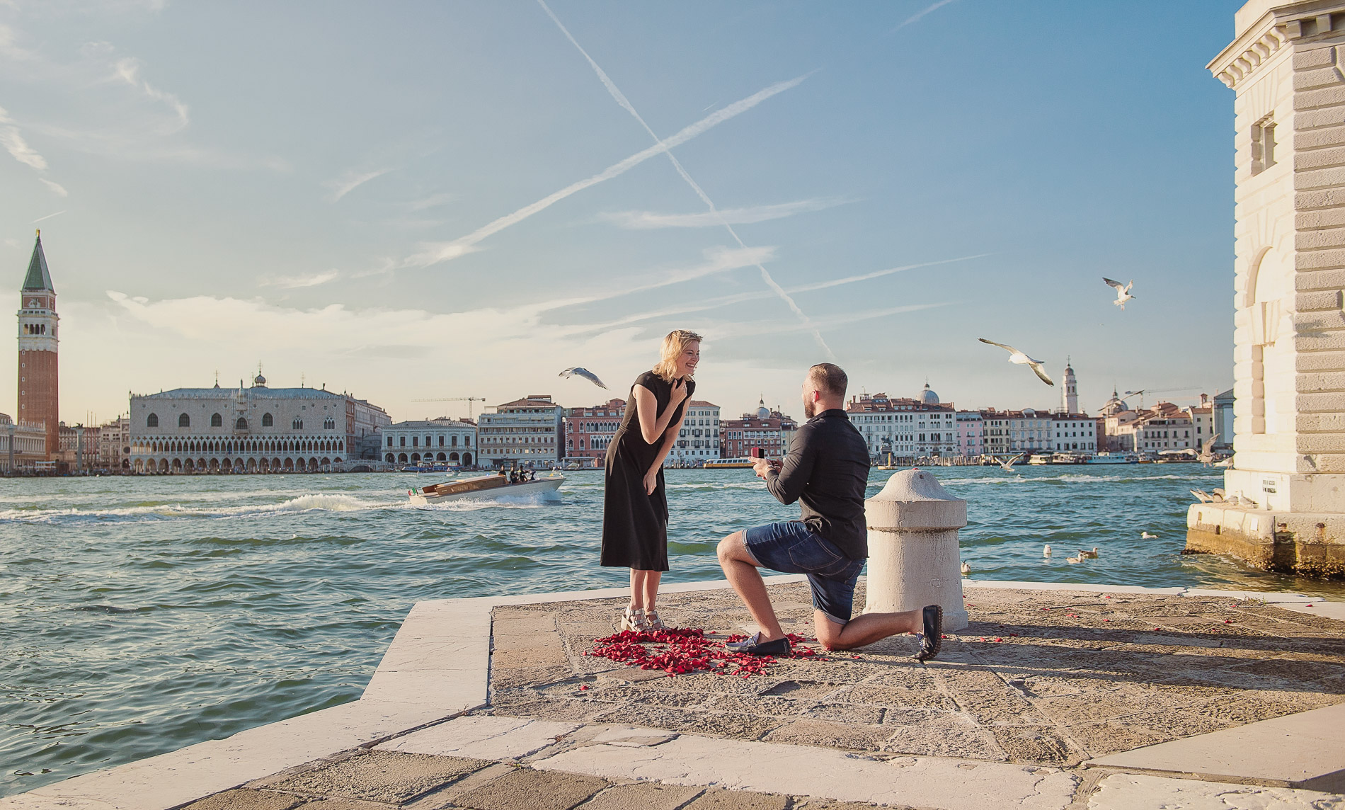 Man on one knee proposing on the hexagonal promontory of San Giorgio Maggiore island, bright open sky, Venice lagoon and city skyline visible in the far background