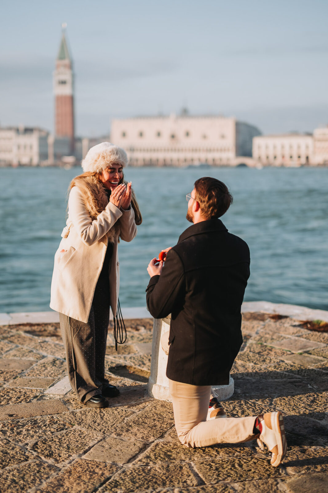Couple embracing on the Venice waterfront, man in dark coat, warm golden light at sunset during proposal photography session