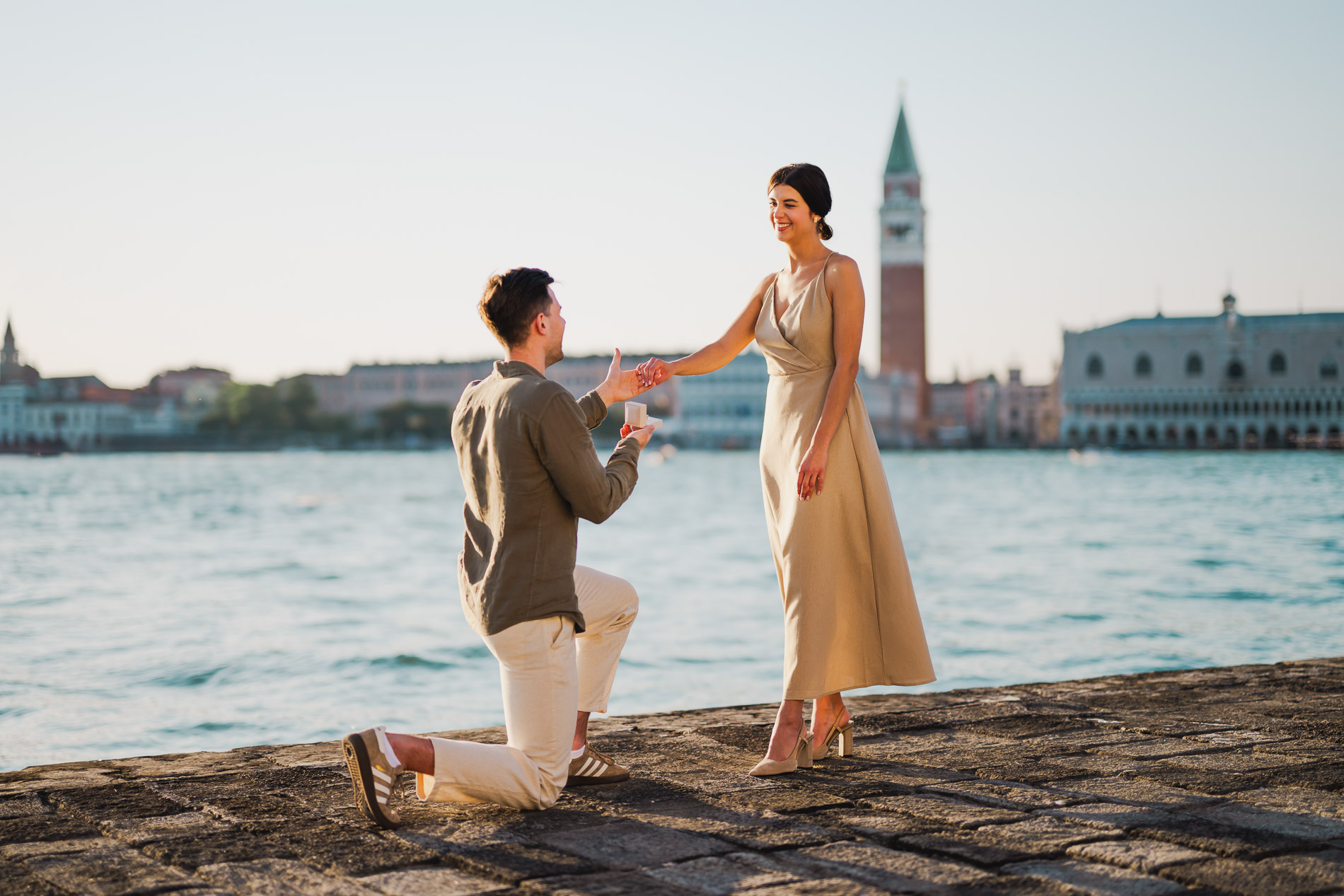 Man on one knee proposing to his partner on the Venice waterfront, woman reacting with emotion, surprise proposal photography session at San Giorgio Maggiore in Venice