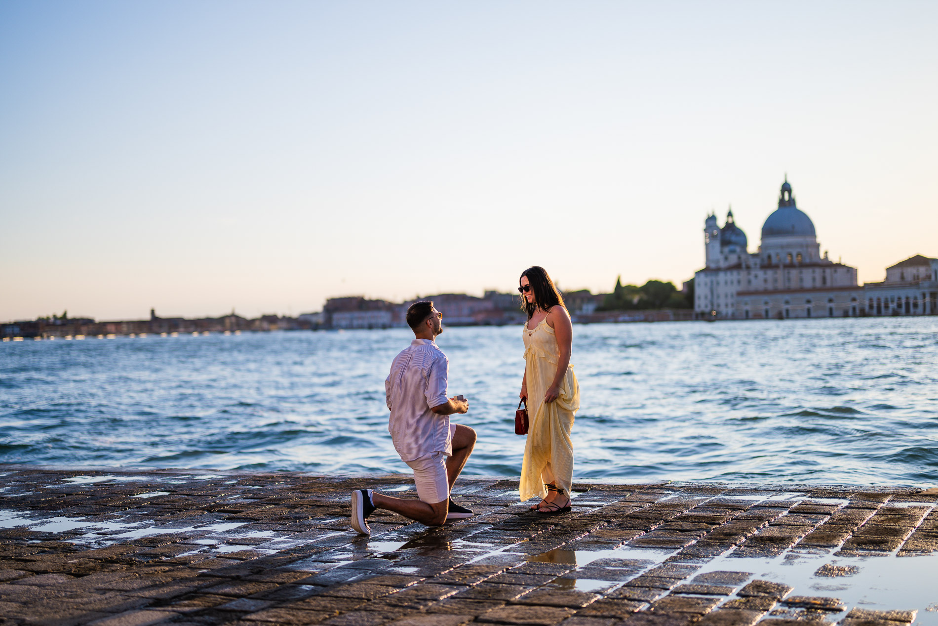 Man going down on one knee to propose on the San Giorgio Maggiore island waterfront, woman reacting, Santa Maria della Salute and Venice skyline in the background