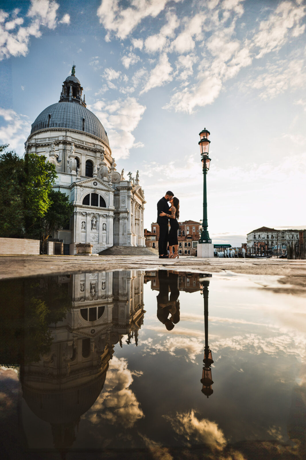 Romantic couple proposing near Venice's historic church at sunset with reflections in water.