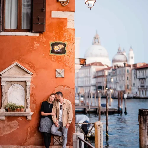 Romantic Venice couple photo by canal, colorful architecture, iconic domes in background.