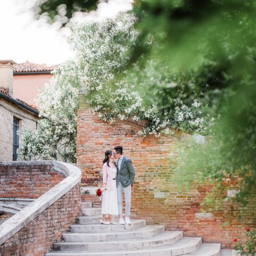engagement-photography-portfolio-in-venice–29 A couple standing on a Venetian staircase surrounded by lush greenery and blooming trees, capturing a romantic atmospher.